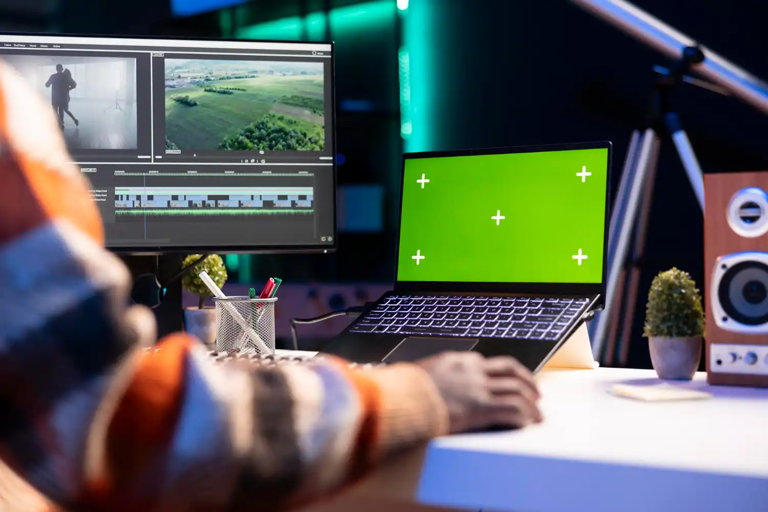Pessoa trabalhando em um computador com tela de edição de vídeo, ao lado de um laptop com tela verde, em um ambiente criativo de produção de conteúdo.